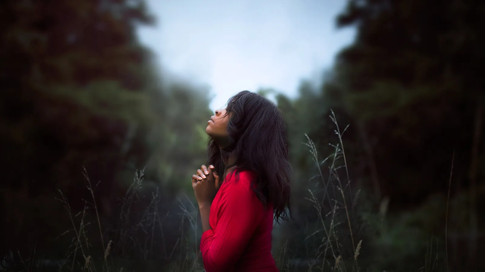 A young Black woman in a red top standing in tall grass with her hands clasped, looking up at the sky in quiet reflection