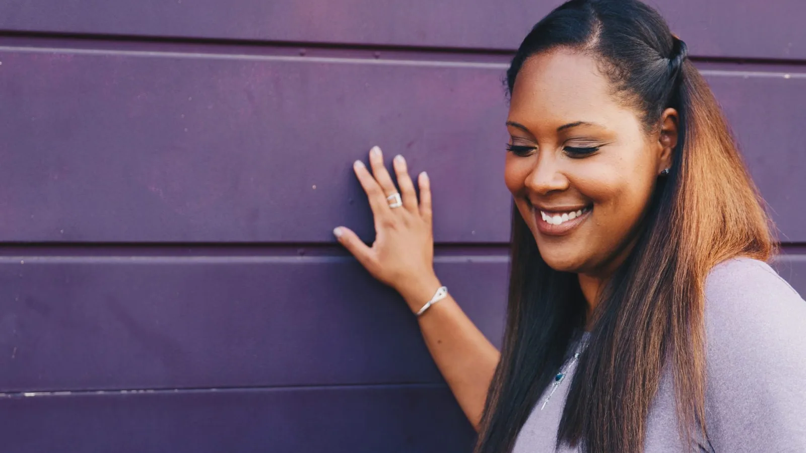 A smiling Black woman resting one hand on a purple wall, looking down with a relaxed, happy expression