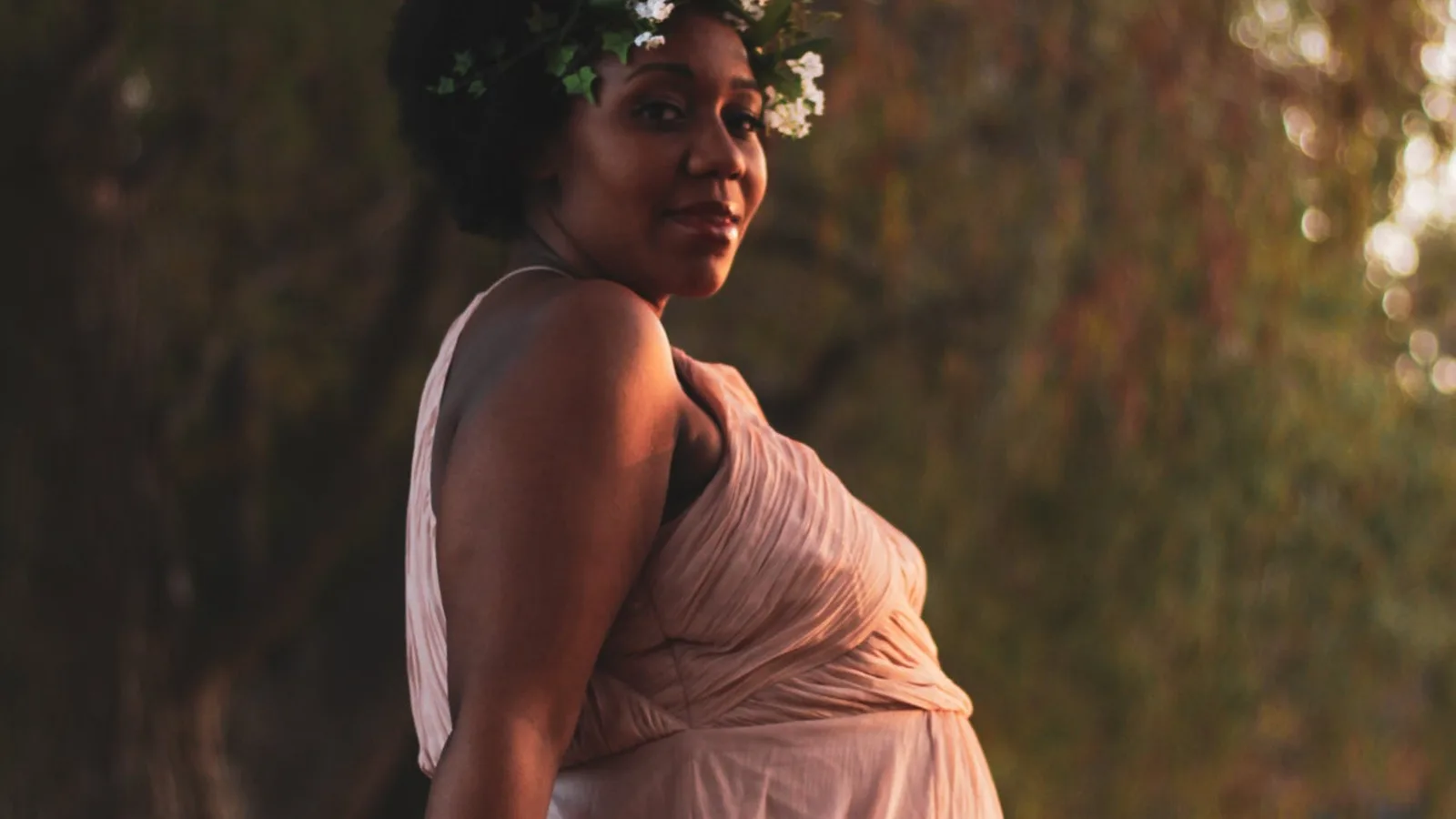A pregnant Black woman in a blush pink gown with flowers in her hair, glancing back over her shoulder in warm evening light